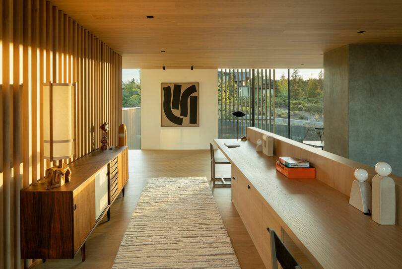 A modern living space with wood accents, a long desk, a sideboard, abstract wall art, and large windows overlooking greenery.
