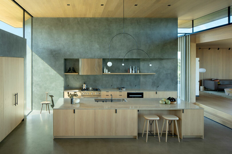 Modern kitchen with light wood cabinets, concrete island, minimalist decor, and built-in appliances. Open shelves display ceramics; pendant lights hang above island with two stools.