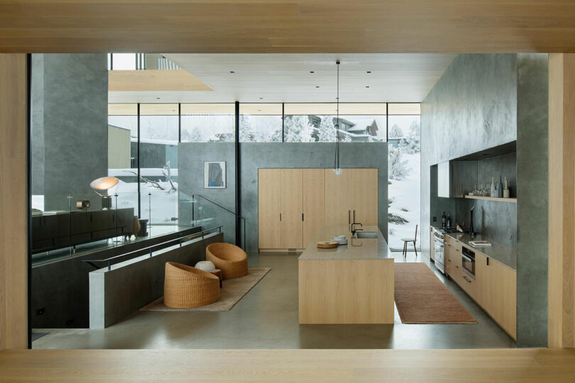 Modern open-concept kitchen and living area with minimalist design, wood accents, two woven chairs, an island, and large windows overlooking a snowy outdoor scene.