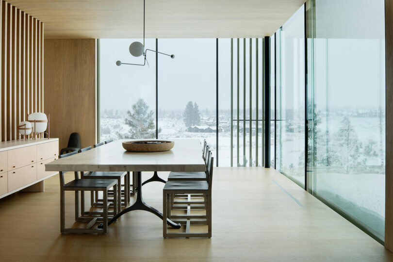 Modern dining room with a large rectangular table, eight chairs, minimalist decor, wood floors, and floor-to-ceiling windows overlooking a snowy landscape.