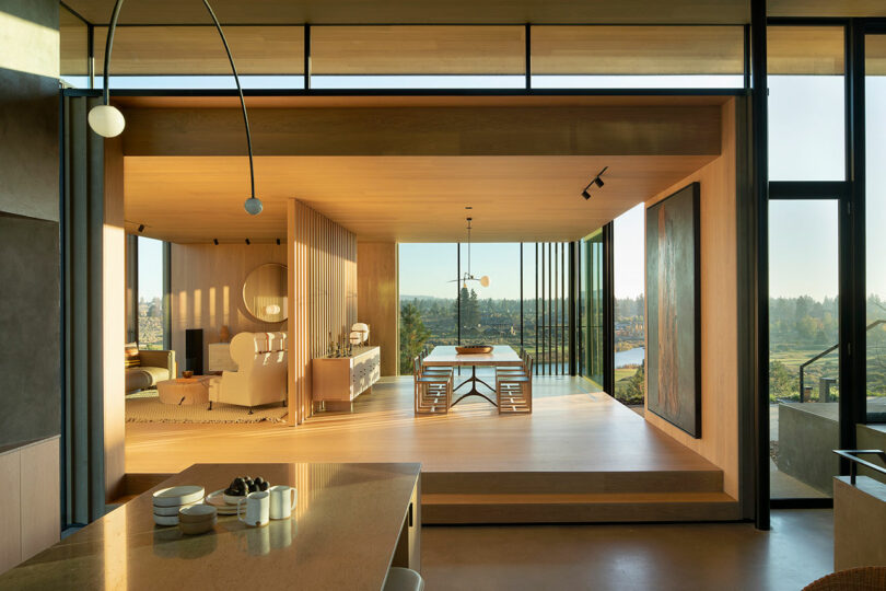 Modern open-plan interior with living and dining areas featuring wood finishes, large windows, and natural light; scenic outdoor view in the background.