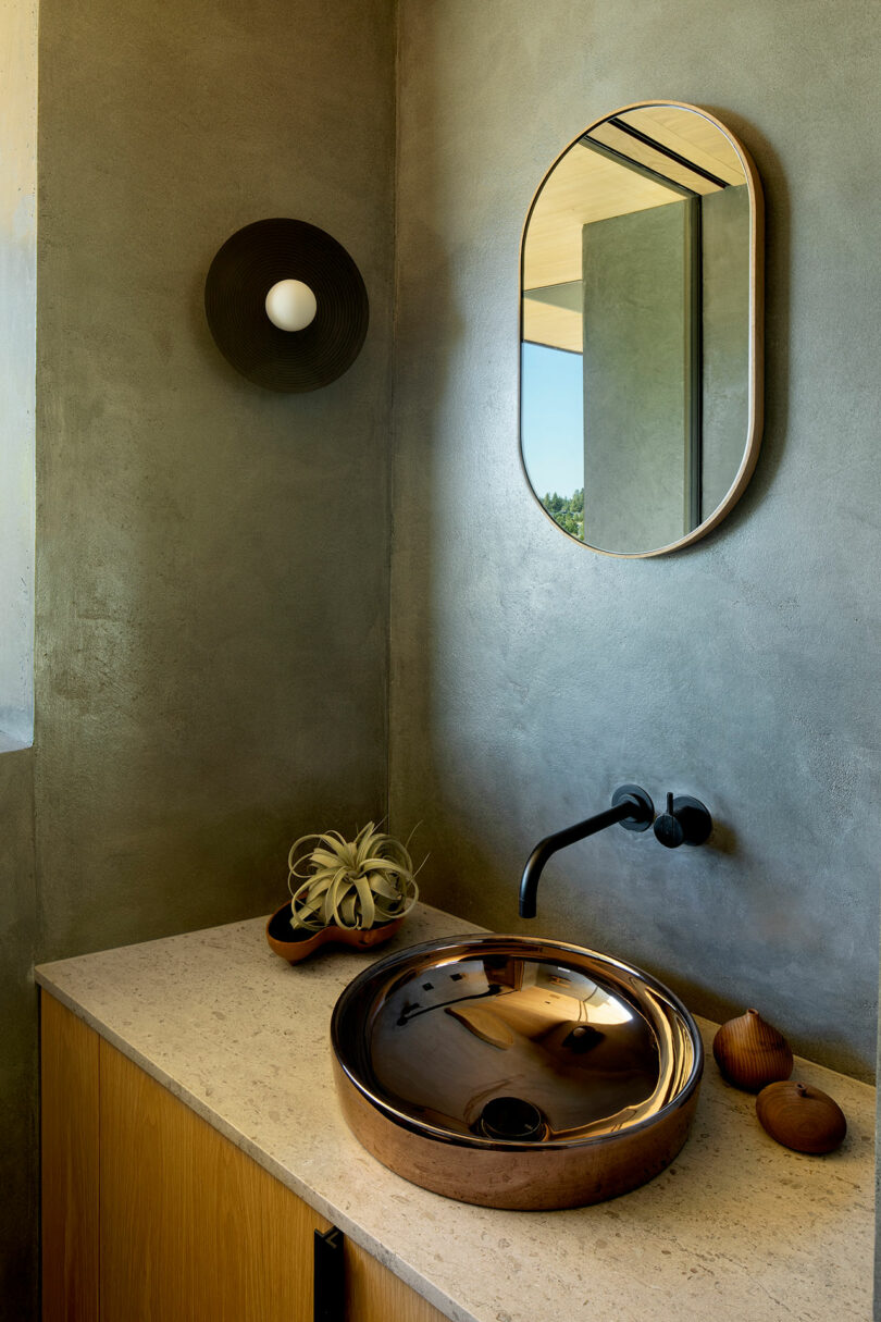 A modern bathroom features a round metallic sink, a wall-mounted faucet, an oval mirror, a black sconce, and small decorative items on a beige countertop with gray walls.