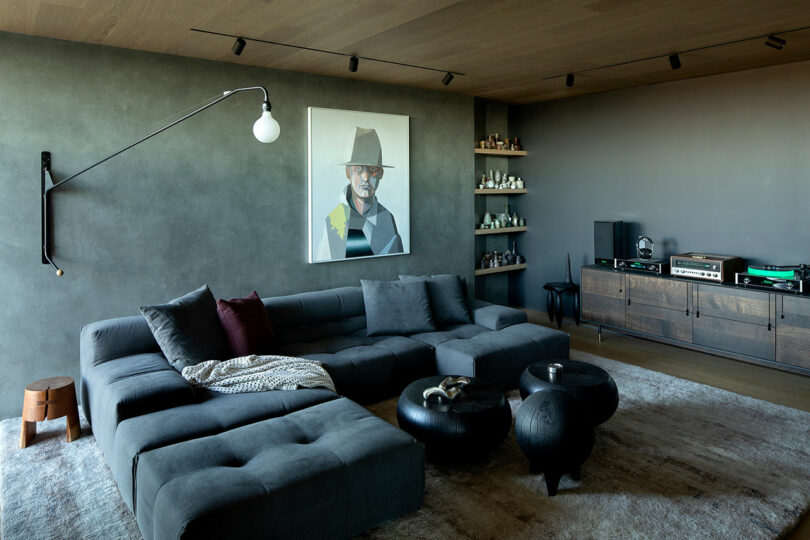Modern living room with a gray sectional sofa, wall-mounted lamp, abstract portrait, shelves with decor items, and a cabinet holding audio equipment and turntables.