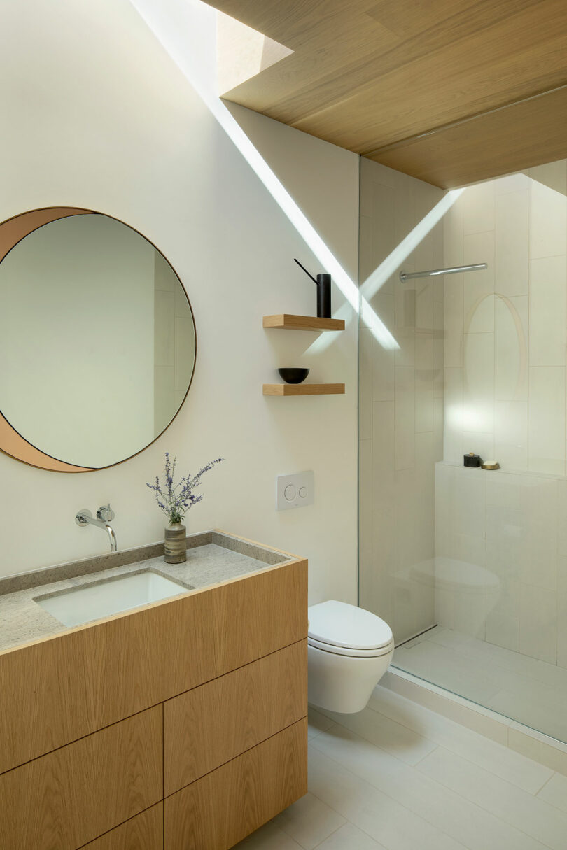 Modern bathroom with a round mirror, wooden vanity, wall-mounted sink, two floating shelves, toilet, and glass-enclosed shower under a skylight.