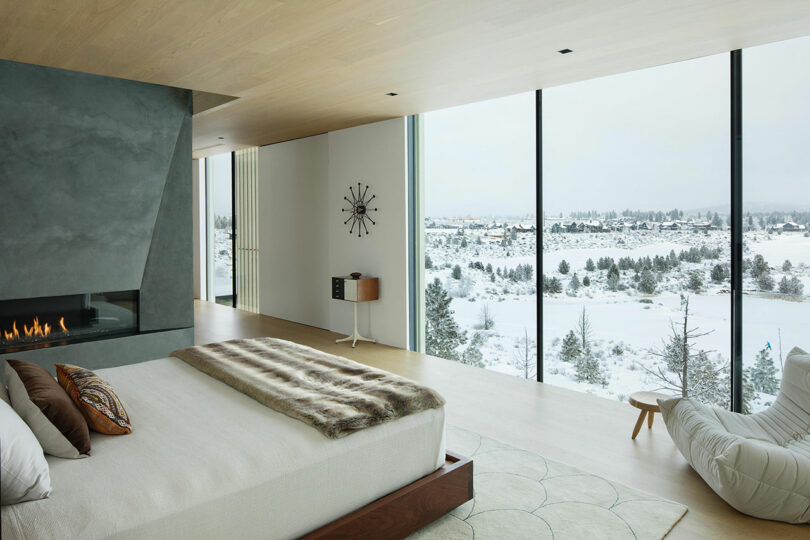 Modern bedroom with large floor-to-ceiling windows overlooking a snowy landscape, a fireplace, and minimalist furnishings including a bed with a fur throw.