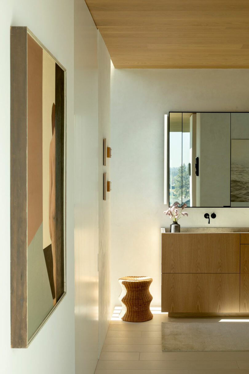 A modern bathroom with wood cabinetry, a lighted mirror, a small patterned stool, and abstract art on a wall.
