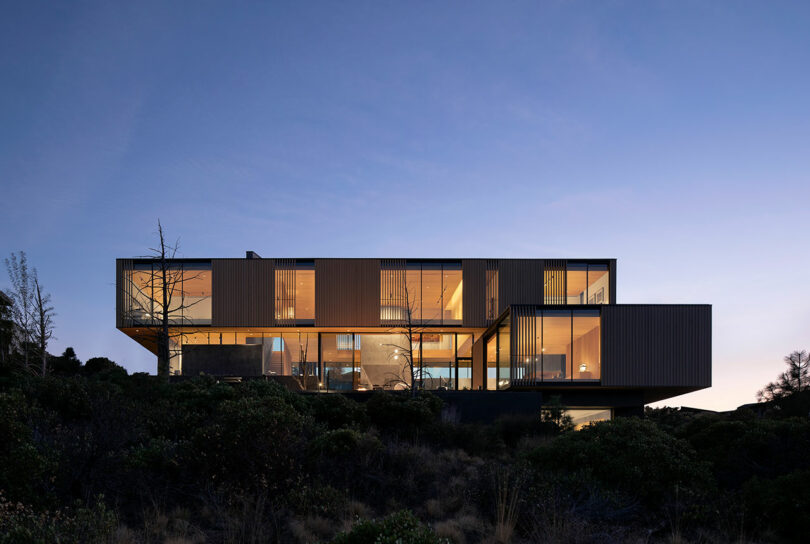 Modern, boxy two-story house with large glass windows, illuminated from within at dusk, surrounded by shrubs and trees.