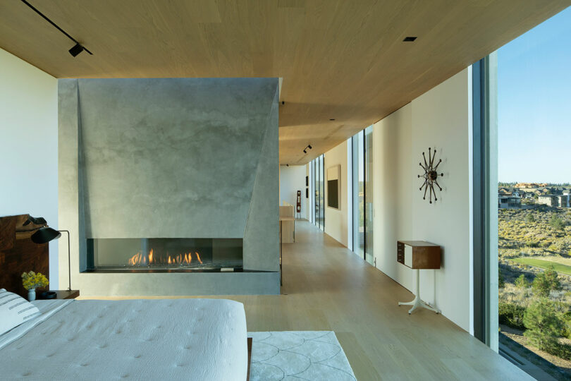 Modern bedroom with a concrete fireplace, light wood floors and ceiling, large windows with views, and minimalistic decor.