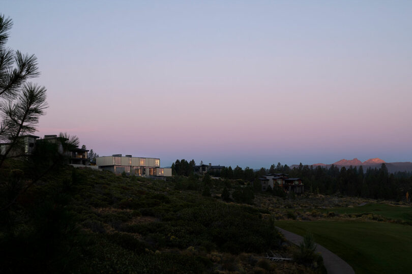 Modern houses sit on a hillside at dusk, with a view of distant mountains and a golf course, under a clear pastel sky.