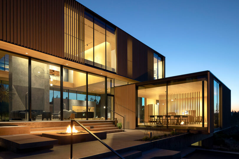 Modern two-story house with large glass windows, wood accents, and exterior lighting, viewed at dusk. A lit fire pit is on the patio in the foreground.