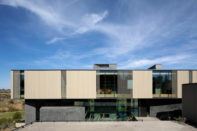 Modern building with large glass windows, vertical beige panels, and concrete base under a blue sky with scattered clouds.