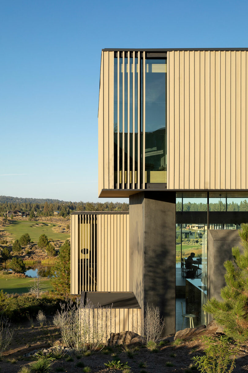 Modern two-story house with vertical wooden slats and large glass windows, situated in a landscaped area with distant trees and hills under a clear sky.