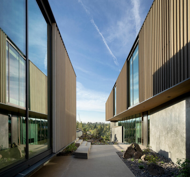Two modern buildings with large glass windows and vertical wooden paneling face each other, separated by a paved walkway with benches and rocks, under a clear blue sky.