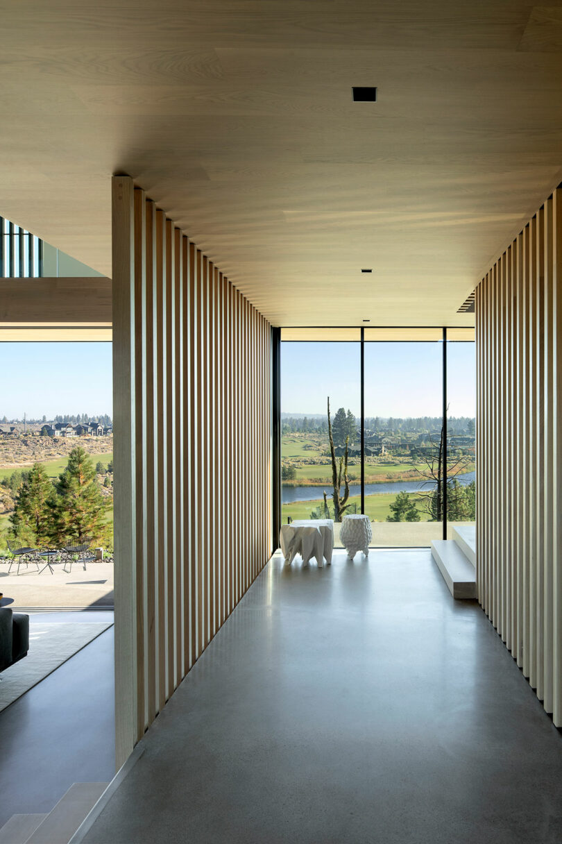 A modern hallway with vertical wooden slats on both sides leads to large floor-to-ceiling windows, showcasing a scenic outdoor landscape with hills and trees.