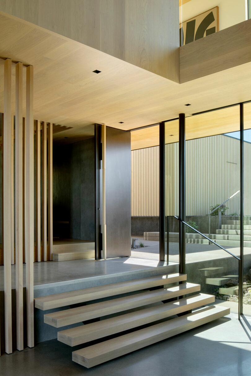 Modern interior with light wood stairs, vertical wooden slats, large glass windows, and a minimalist open entrance leading to an outdoor area.