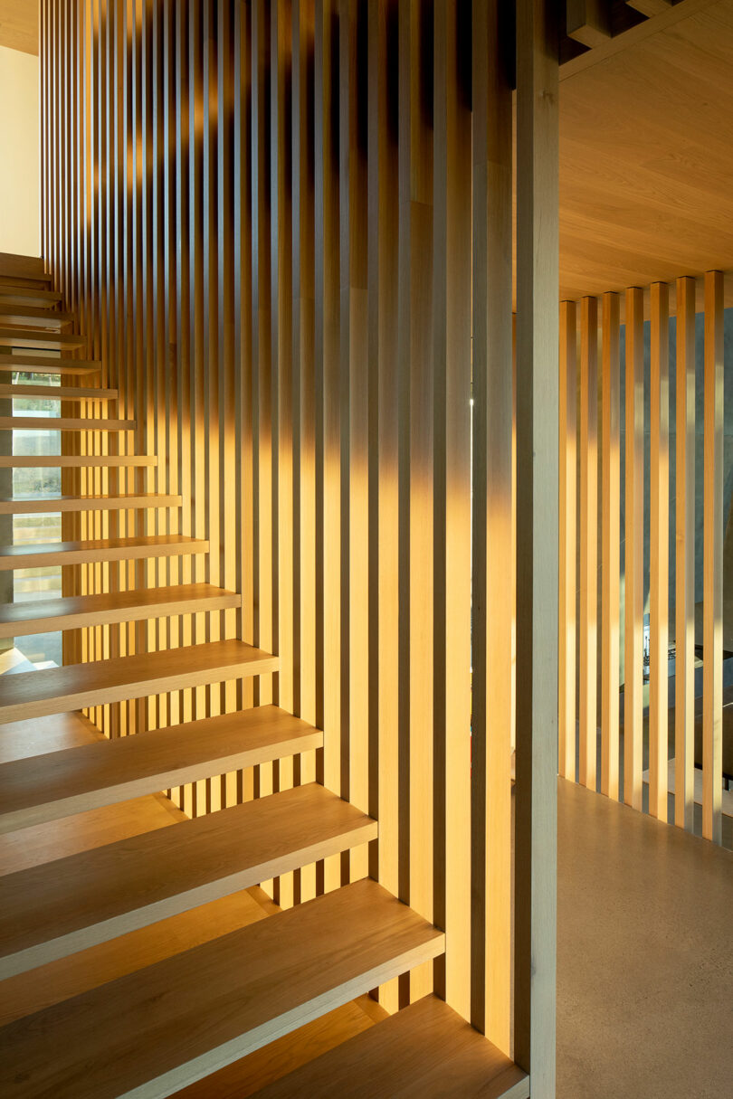 Floating wooden staircase with vertical wooden slats, casting shadows on the wall and floor in a modern interior space with warm lighting.