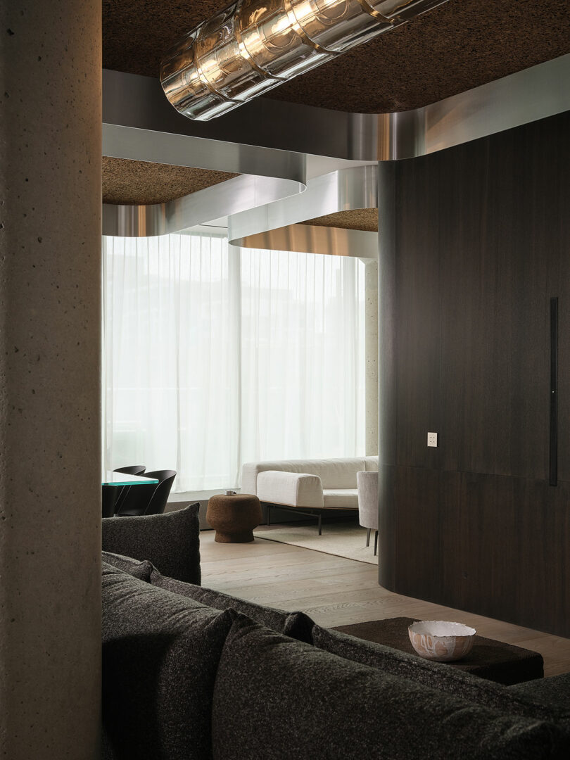 Modern living room with dark wood paneling, a concrete pillar, sheer curtains, black and white furniture, and a metallic ceiling fixture. Natural light fills the space.