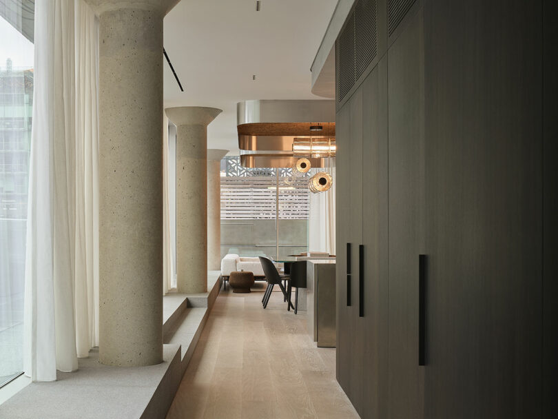 Modern interior with large columns, floor-to-ceiling windows, light wood flooring, dark cabinetry, and a dining area with contemporary lighting fixtures.