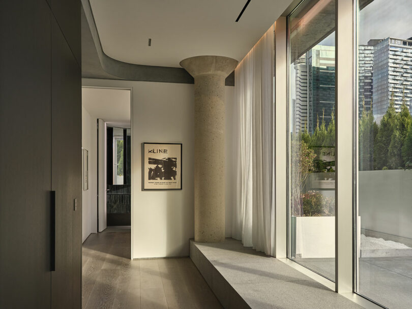A modern interior hallway with a concrete pillar, large window, sheer curtains, wood flooring, and a framed poster on the wall; city buildings visible outside.