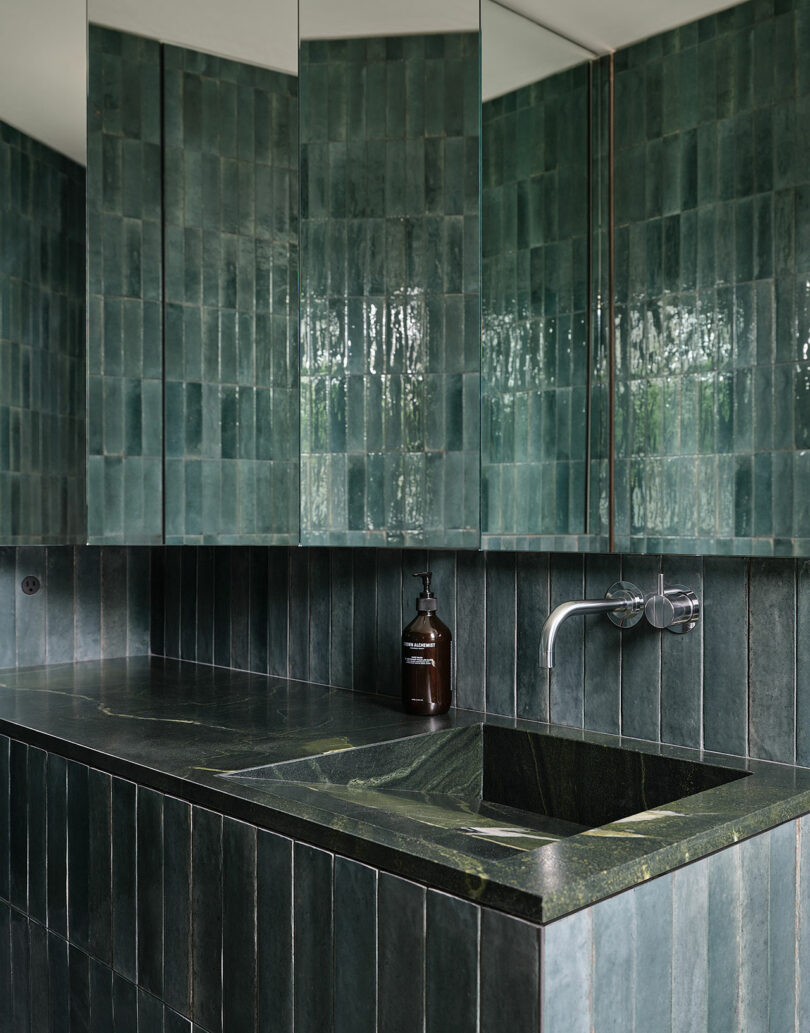 A modern bathroom with green vertical tiles, a matching countertop and sink, wall-mounted faucet, and a brown soap dispenser.