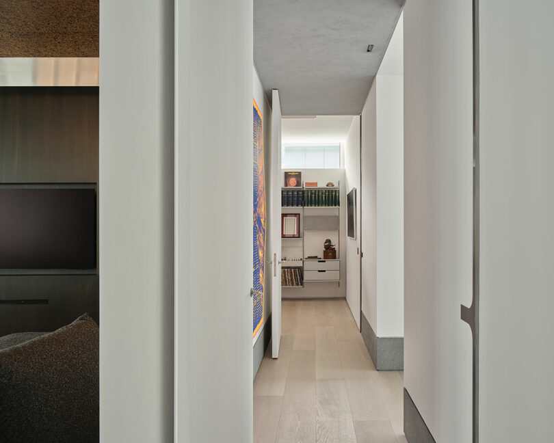 A narrow hallway with light wood floors leads to a small office area with shelves, books, and a desk under a window with natural light.