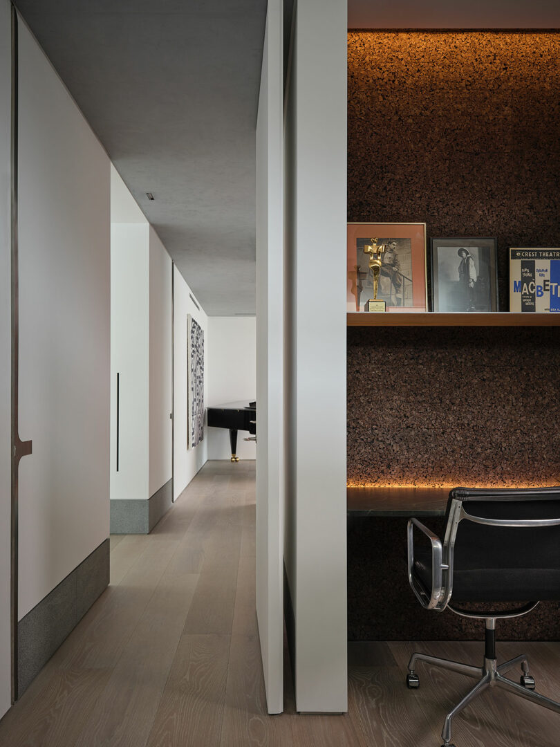A modern hallway with light wood flooring leads to a room with a piano; a built-in desk with shelves, chair, framed photos, and awards is visible on the right.