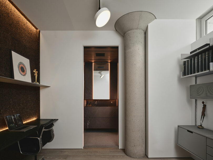Modern office with a desk and shelves on the left, a large concrete pillar near the center, and an open doorway revealing a bathroom with a lit mirror in the background.