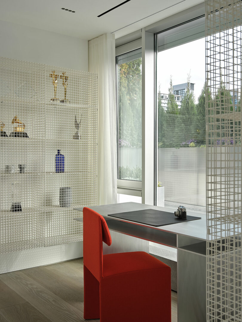 A modern office with a red chair, metal desk, and grid-patterned shelves displaying books and awards, next to large windows overlooking greenery.