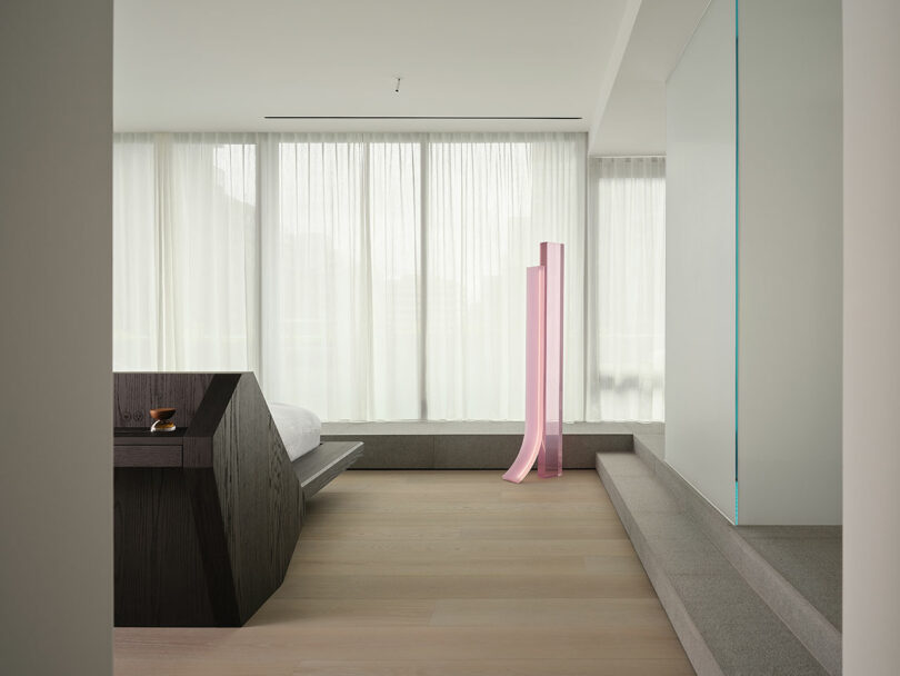 Minimalist bedroom with a low dark wood bed, large sheer curtains, light wood floors, and a tall pink modern sculpture near a raised platform.