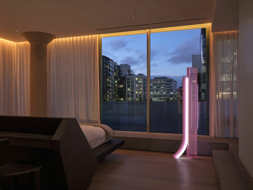 A modern bedroom with floor-to-ceiling window, cityscape view at dusk, sheer curtains, and a tall, pink LED floor lamp beside the bed.