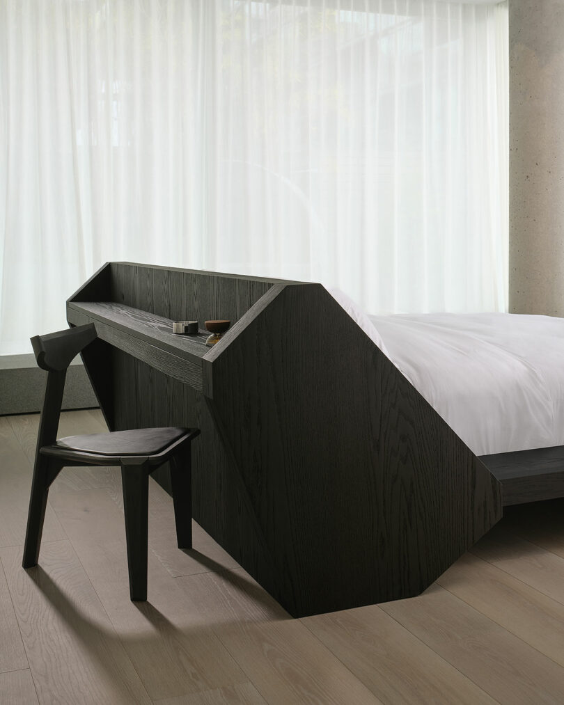 A modern, minimalist bedroom with a geometric dark wood desk attached to the bed frame, paired with a matching chair, light wood floors, and sheer curtains in the background.
