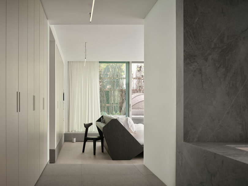 Minimalist living space with neutral tones, featuring a dark angular sofa, black chair, floor-to-ceiling windows with white curtains, and built-in storage cabinets.