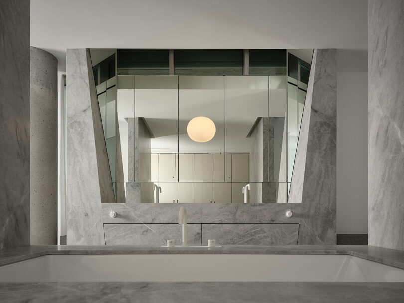 Modern bathroom with grey marble surfaces, a white bathtub, double faucets, large angular mirrors, and a round ceiling light reflected above.