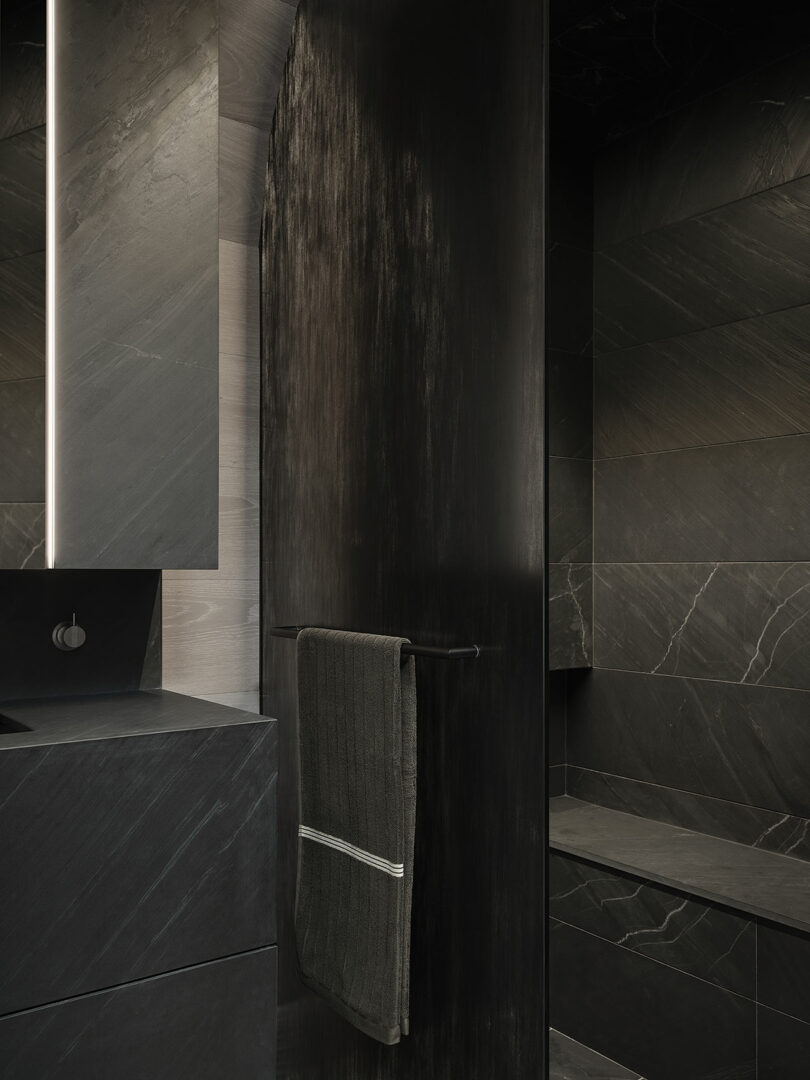 A modern bathroom with dark stone walls, a built-in bench, a towel hanging on a black metal bar, and a tall, illuminated mirror.