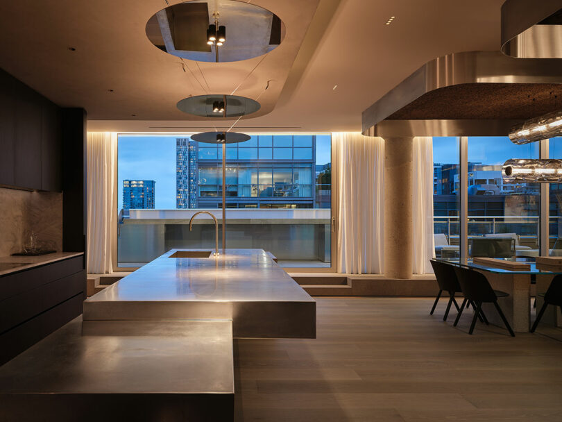 Modern kitchen and dining area with a large stainless steel island, minimalist furnishings, and floor-to-ceiling windows overlooking city buildings at dusk.