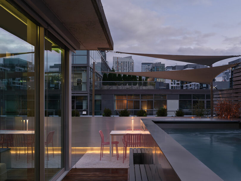 A modern rooftop patio with pink chairs and tables beside a pool, surrounded by city buildings and shade sails, at dusk.