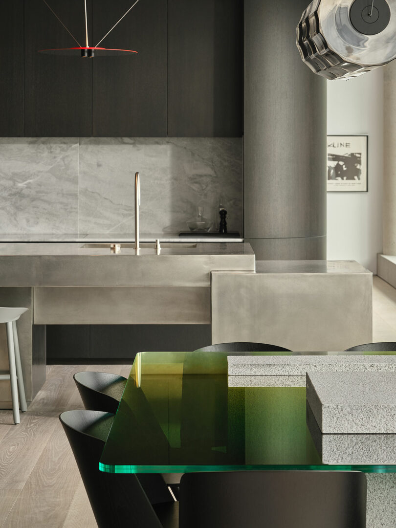 A modern kitchen features a green glass dining table, angular black chairs, marble backsplash, minimalist faucet, and contemporary lighting with neutral tones.