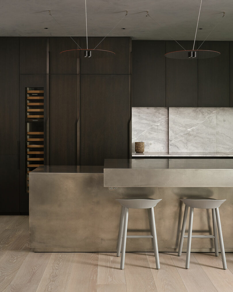 Modern kitchen with dark cabinetry, built-in wine cooler, metallic island, two gray bar stools, and light wood flooring. Marble backsplash and minimalist pendant lights overhead.