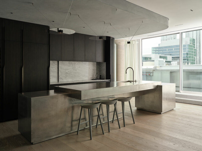 Modern kitchen with minimalist design featuring dark cabinets, a stainless steel island with three stools, marble backsplash, and large window overlooking city buildings.
