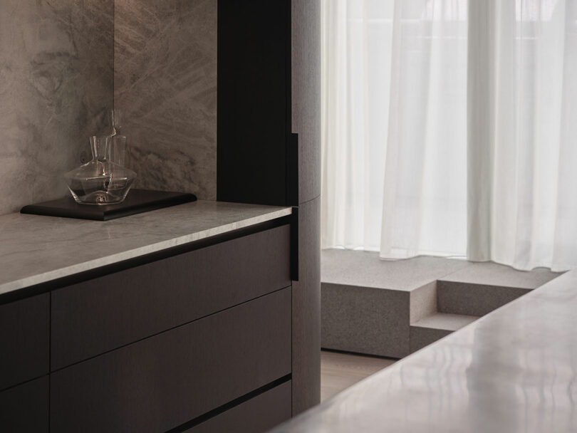 A modern interior with dark cabinetry, a marble countertop, a glass decanter set on a tray, and sheer white curtains letting in natural light.