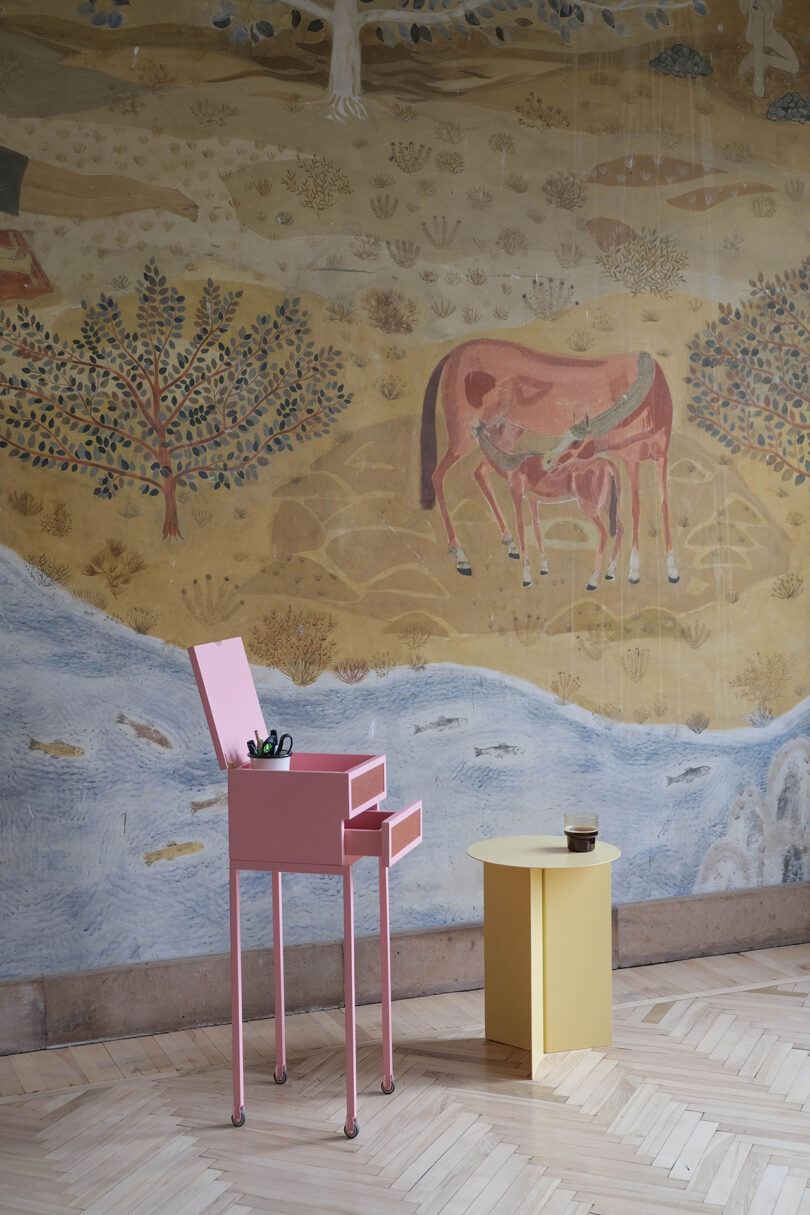 A pink cabinet stands open beside a yellow table on a wooden floor. In the background, a mural depicts cows, trees, and a river.