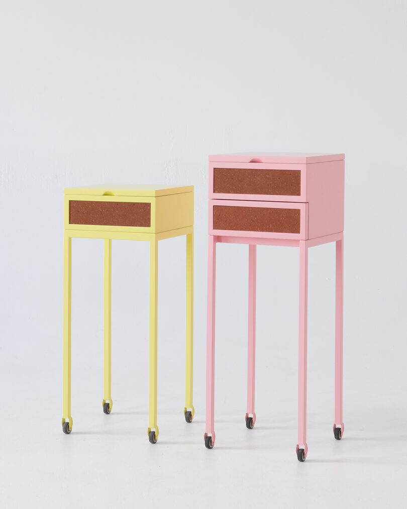 Two colorful side tables on wheels against a white background. One is yellow with one drawer, and the other is pink with two drawers, both featuring brown drawer fronts.