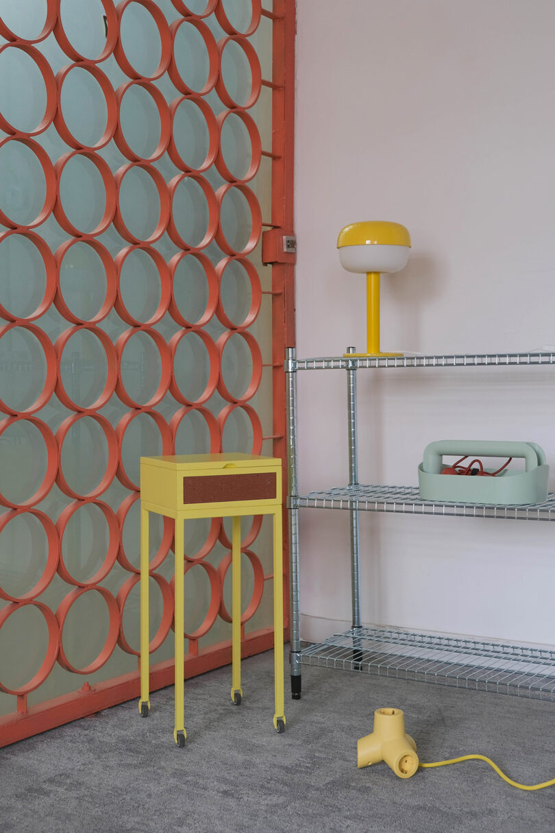 Room with orange circle wall divider, yellow side table, metal shelving unit with yellow lamp, green box, and yellow hairdryer on grey carpet.