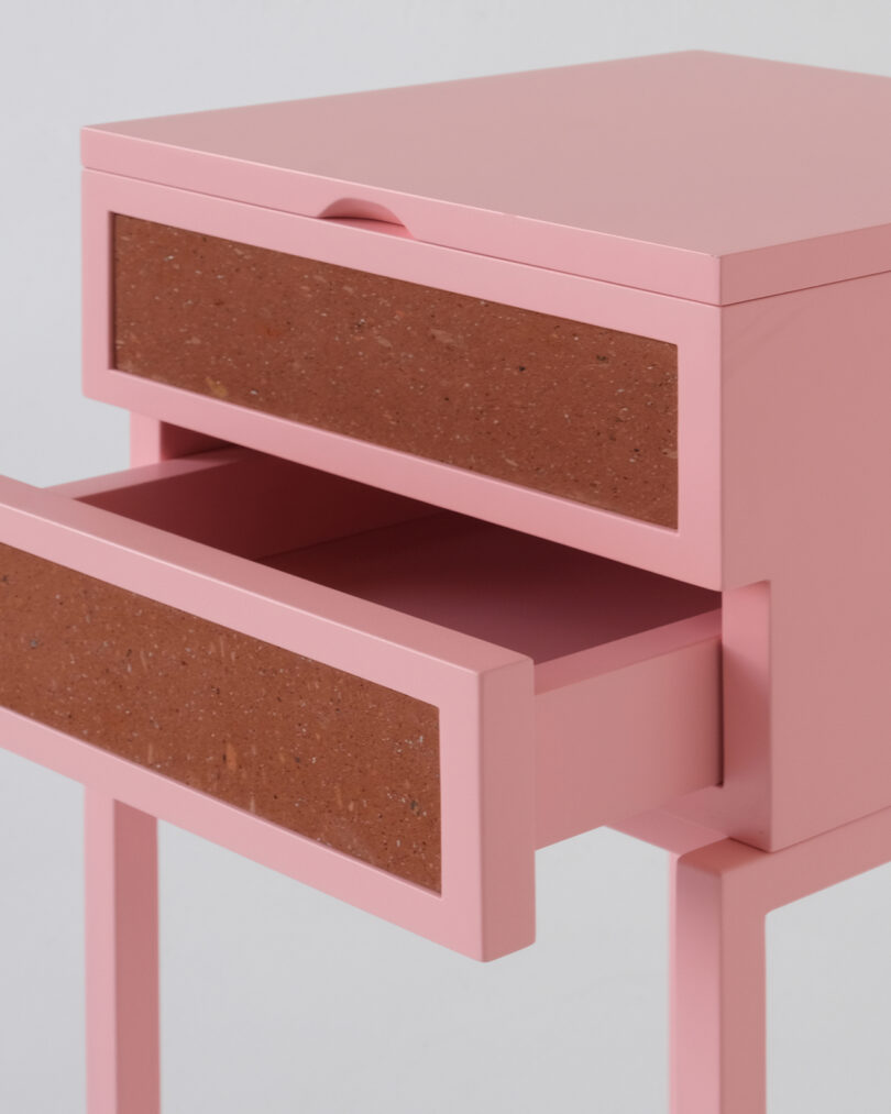 Pink bedside table with two drawers, featuring brick-textured front panels. The lower drawer is partially open.