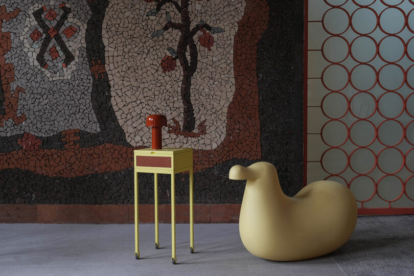 A tall, yellow stand with a small cabinet and red lamp is next to a low, yellow chair. The background features a mosaic wall with abstract patterns and a grid of circular windows.