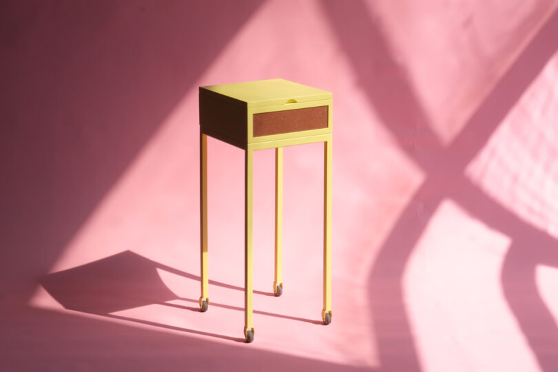 Yellow side table on wheels with a single drawer, placed on a pink background with shadow patterns.
