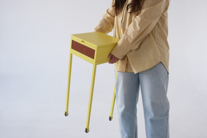 Person in a beige shirt and jeans holding a small yellow side table with wheels against a plain background.