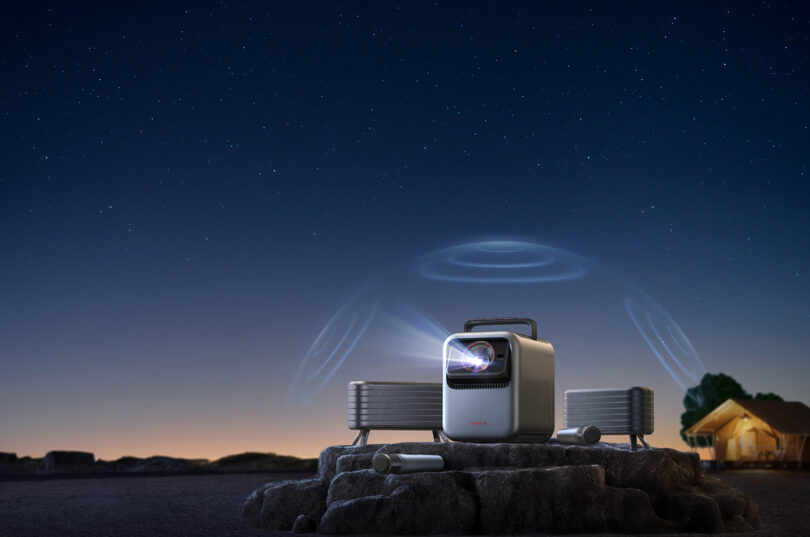 A portable projector is set up on a rocky surface outdoors at night, projecting light, with speakers nearby and a tent visible in the background under a starry sky.