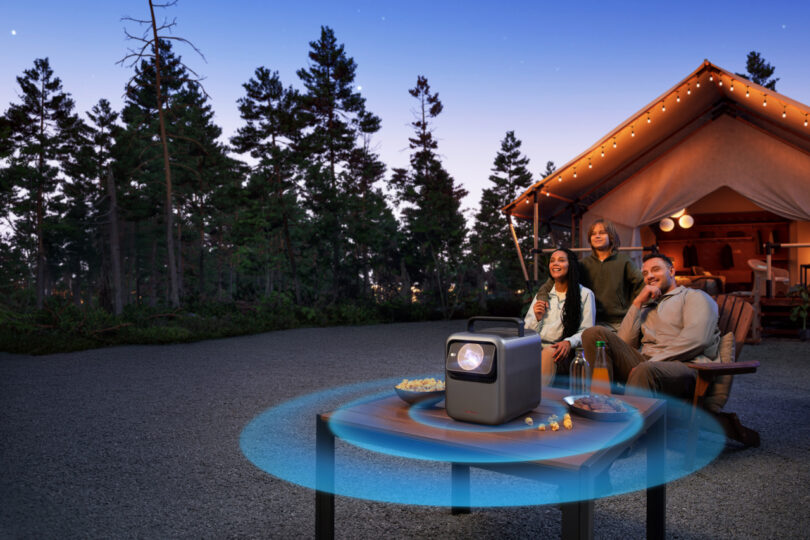 Three people watch a projector outdoors beside a tent at dusk, seated on chairs around a table with snacks and drinks. The projector emits blue sound waves.