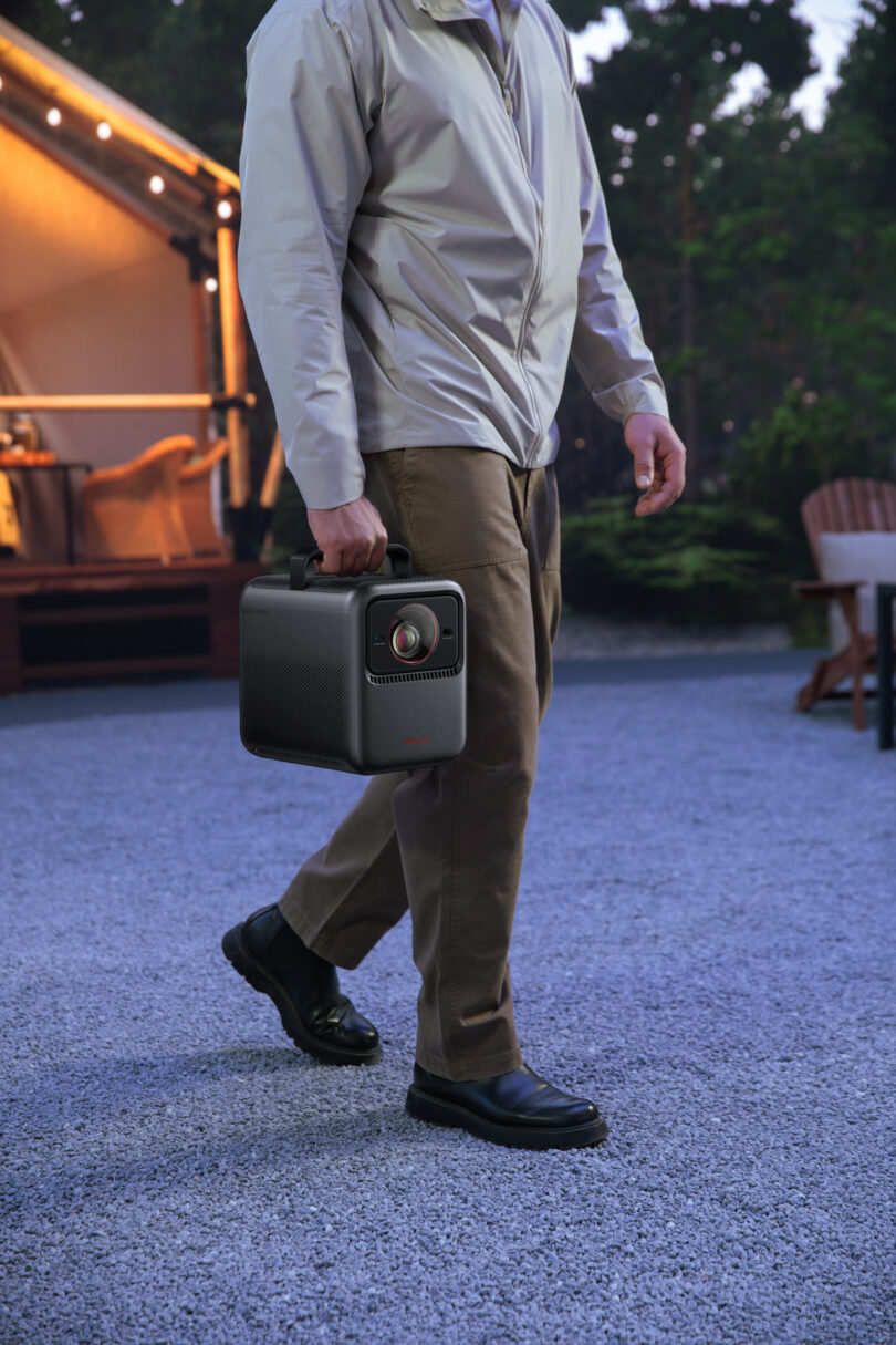 A person wearing a light jacket and brown pants carries a portable projector outdoors on a gravel path near a tent and wooden chairs.